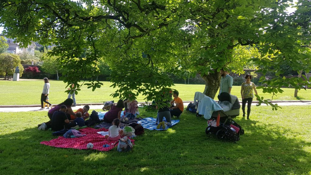 Grupo de familias nun parque, baixo a sombra dunha grande e frondosa árbore e toallas na herba para que estén as crianzas. Día de sol. Primaveira. Tribo
